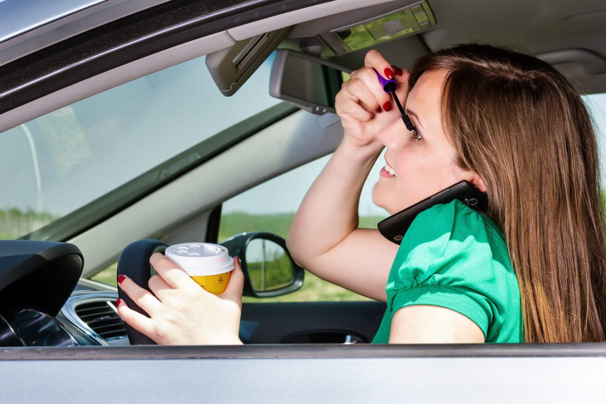 A female wearing a green shirt, sitting in a car, behind a steering wheel, holding both the steering wheel and her coffee in her left hand while looking into the sun visor mirror, putting on mascara.