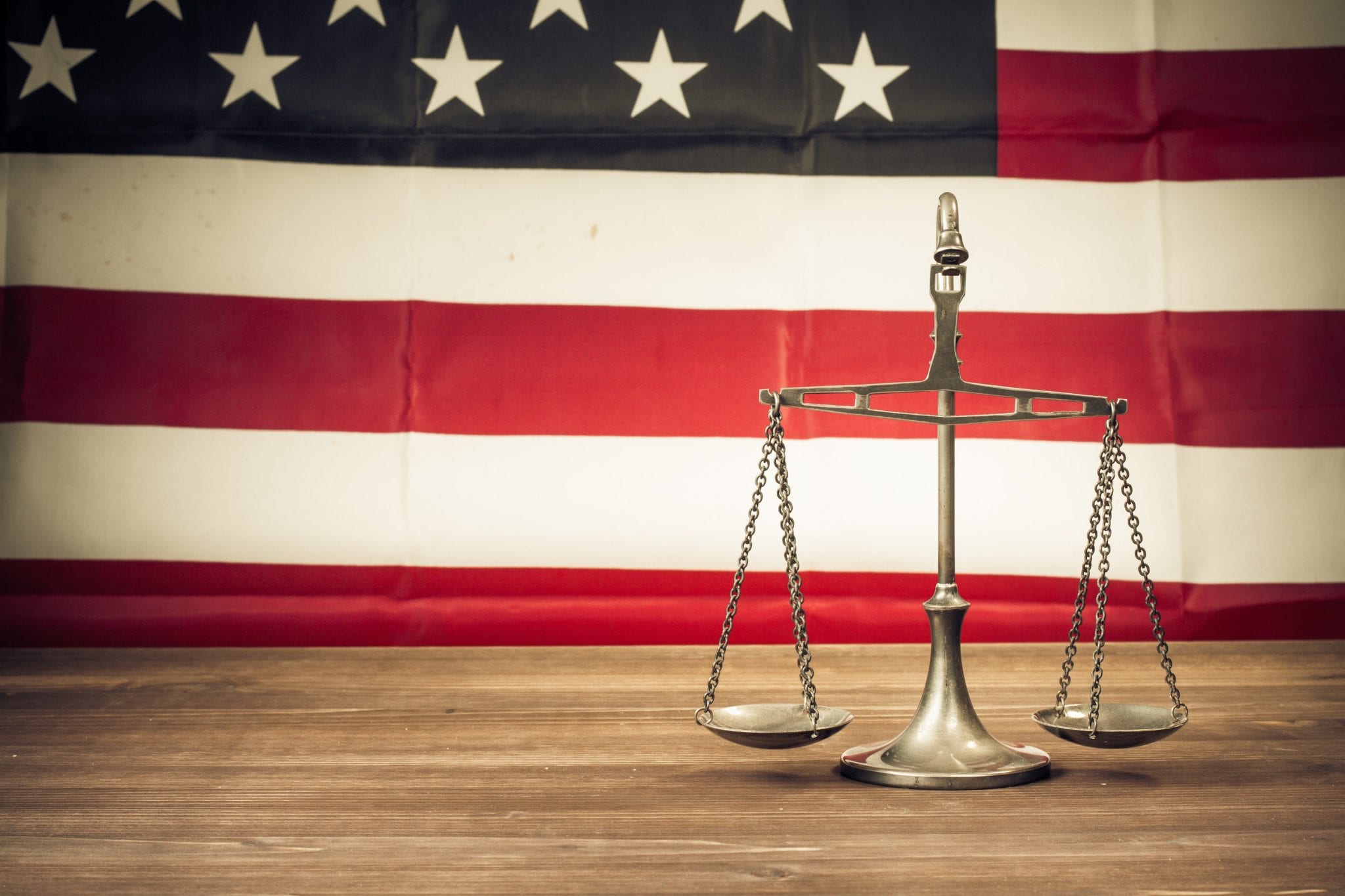 The scales of justice sitting on a wooden table with an american flag background.