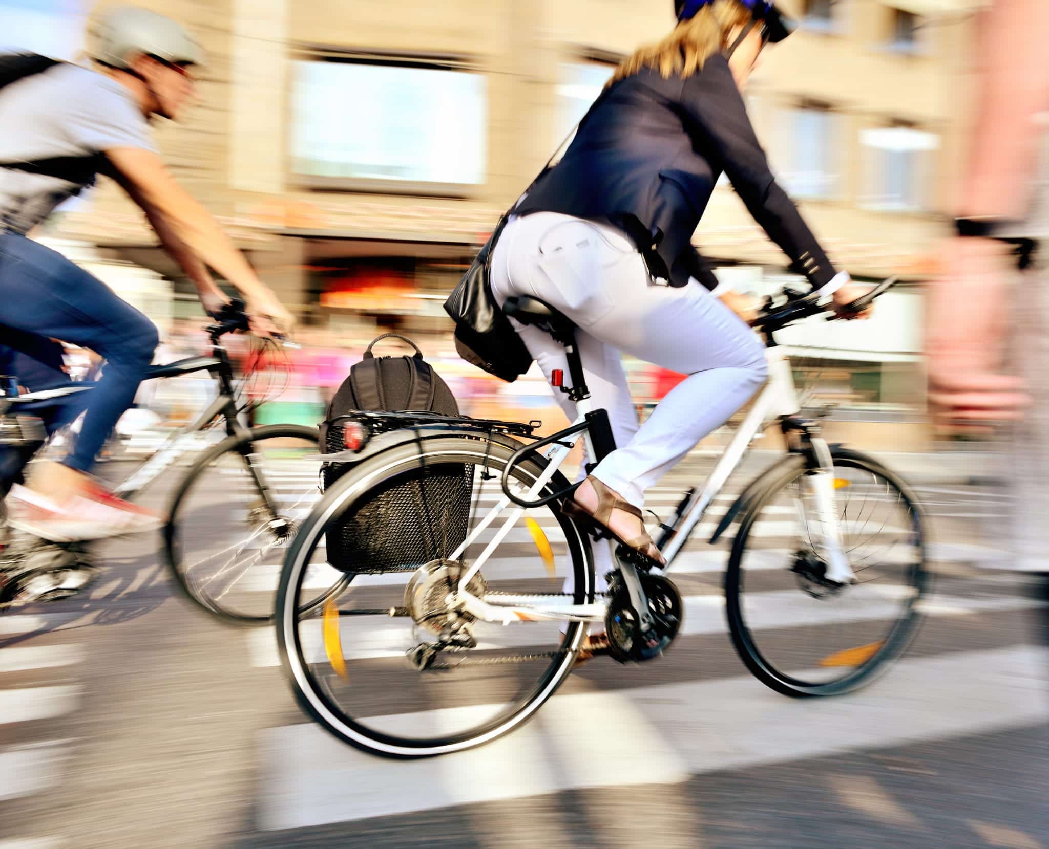 A woman in white jeans and a navy jacket riding a white bicycle through city streets with other cyclists in the blurry background.
