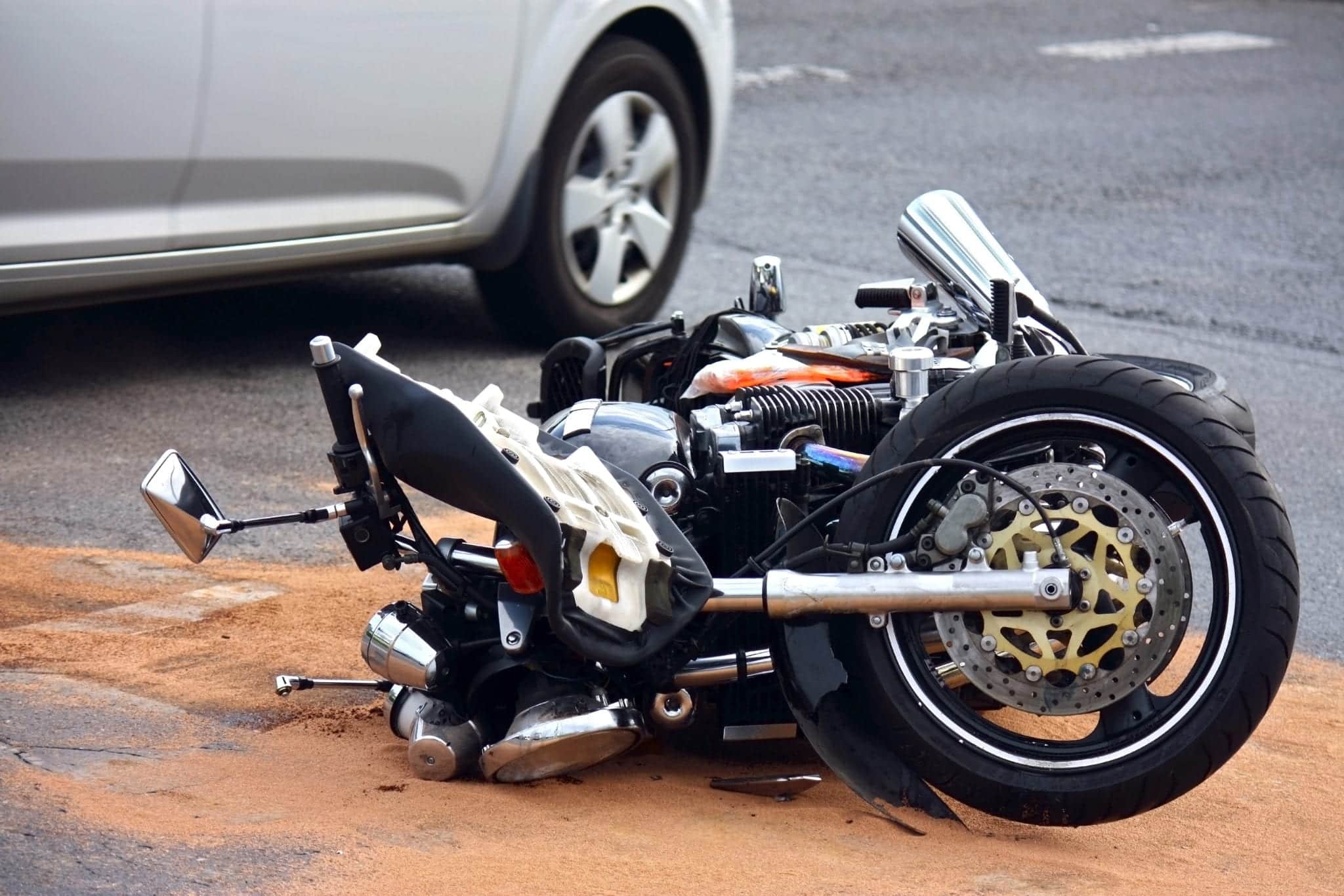 A wrecked motorcycle laid in the road with a silver car in the background.