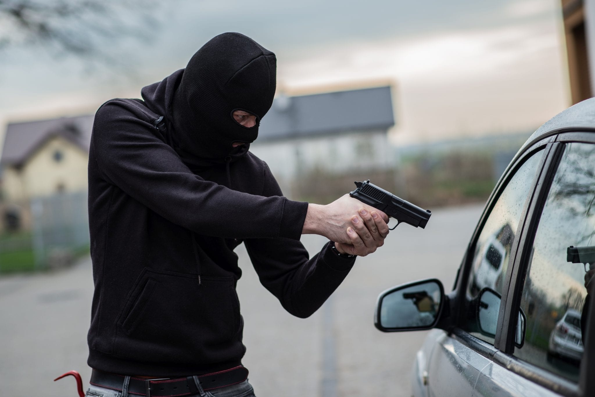 An armed, masked man, wearing all black, pointing a gun at a car window.