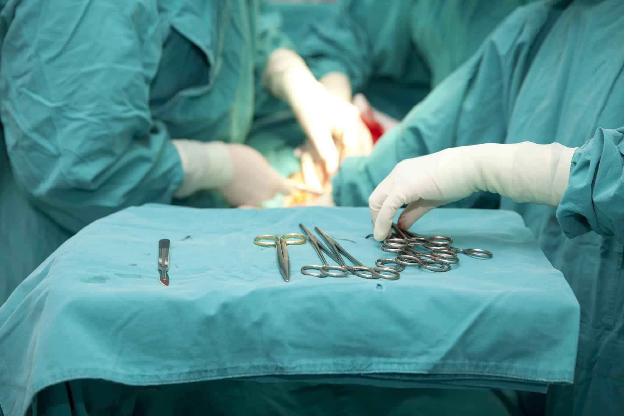 A table covered in a green surgical cloth with a silver surgical tools, a surgical assistants white gloved hand is reaching for a tool and a surgical procedure is being conducted in the blurred background.