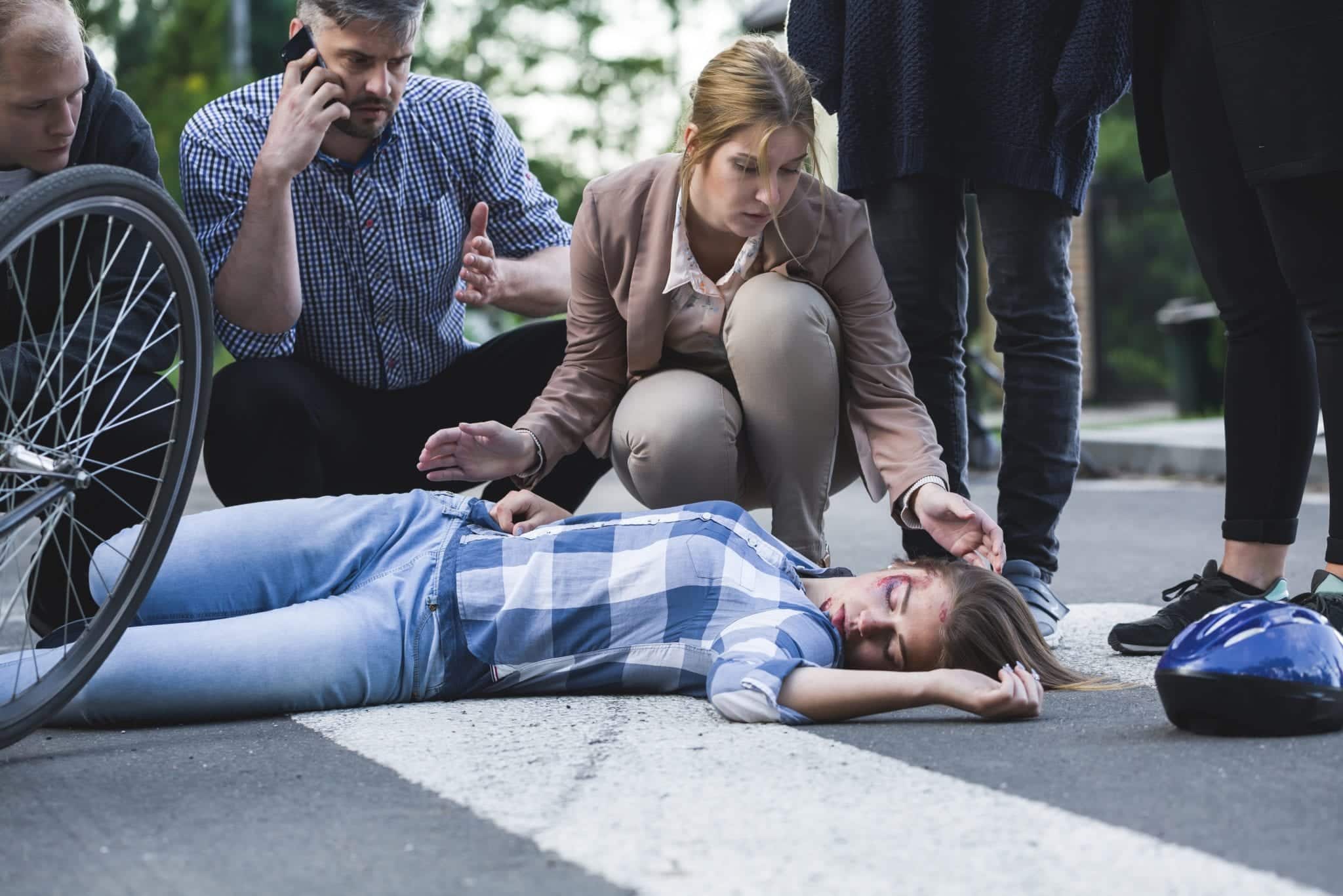 Several people kneeling down, tending to an unconscious woman laying on the concrete beside a bicycle tire with a helmet laying off to the side.